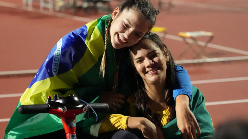 A woman in a wheelchair hugging another woman sitting on the ground
