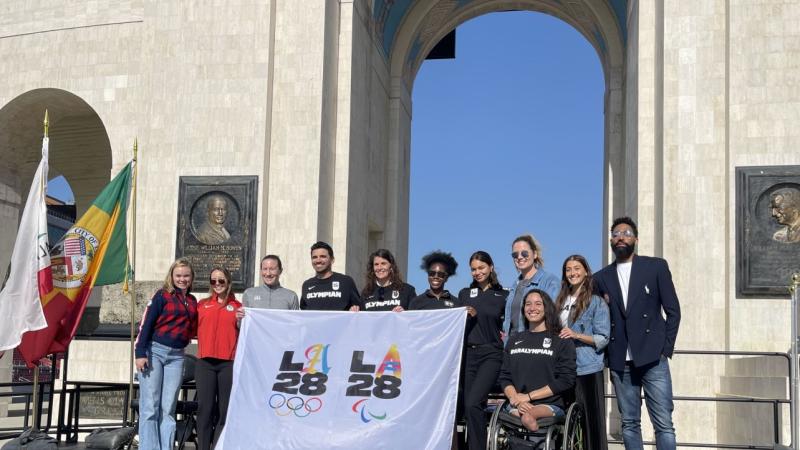 11 people including one female Paralympian pose for a group photo in front of a stadium. They are holding a white flag with the logos of the LA Games