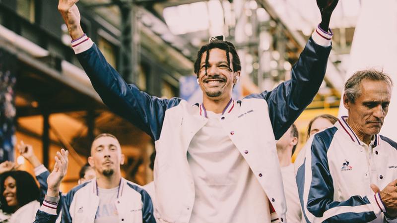 A male athlete wearing a white and blue hoodie is opening his arms wide to the crowd
