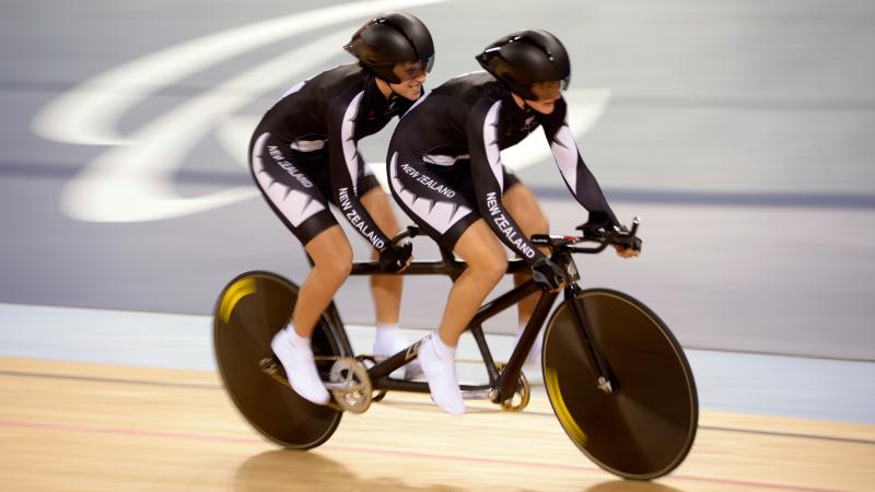Two female cyclists in black racing suits on a tandem, competing at the London 2012 Paralympic Games.