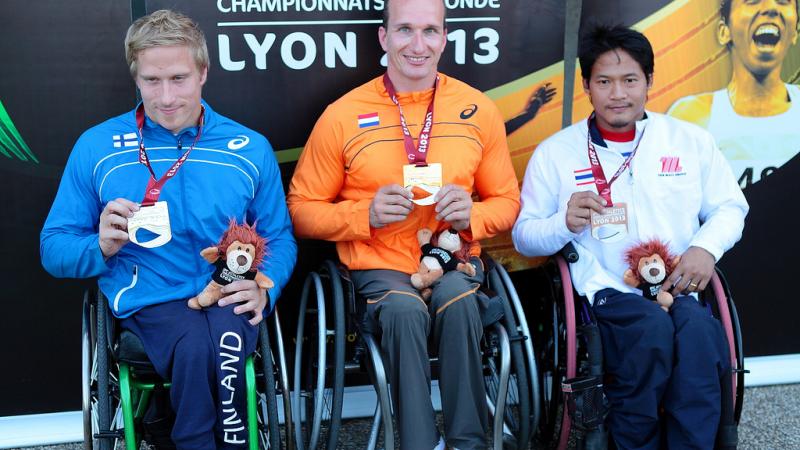 Three wheelchair athletes pose with their medals in front of a Lyon branded background
