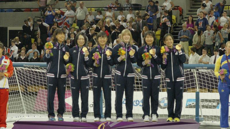 Six women in training suits on podium with medals and flower boquets.