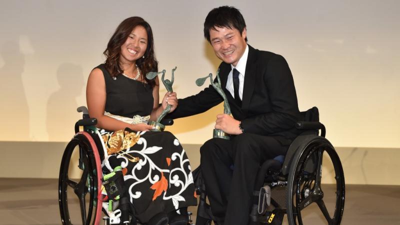 Man and woman in wheechairs holding trophies and smiling to the camera