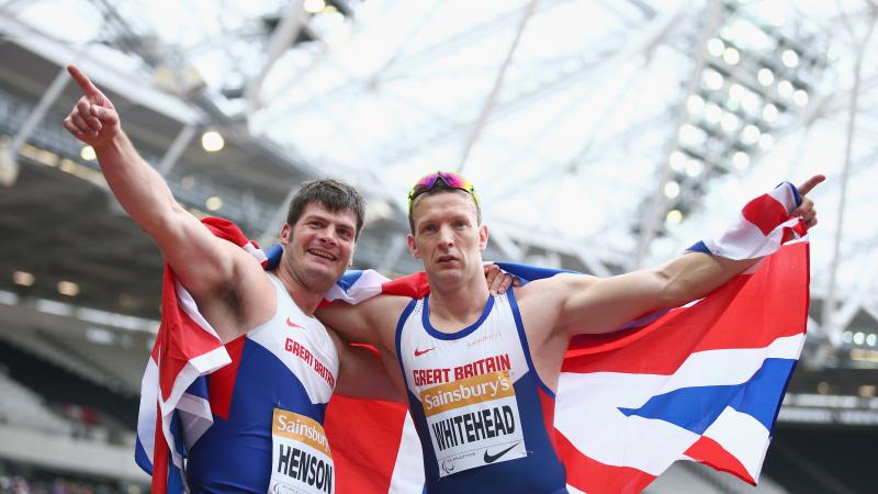 Two men in British racing vests hold a union jack behind them as they celebrate victory,