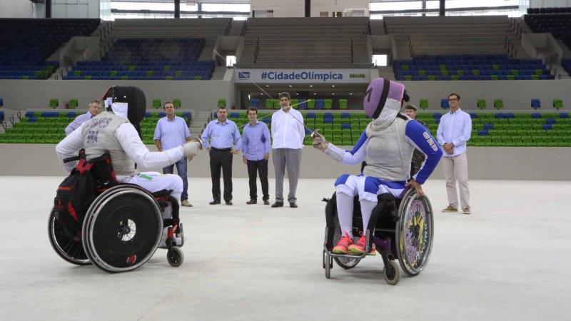 Two wheelchair fencers cross swords in empty arena, some people watching from behind