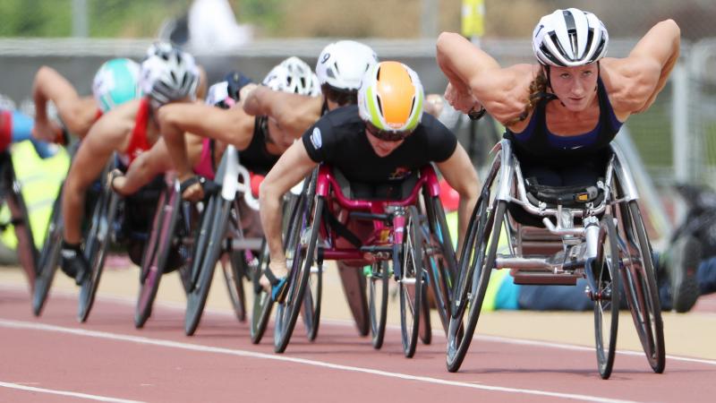 A wheelchair racer leads a line of competitors on a red track.