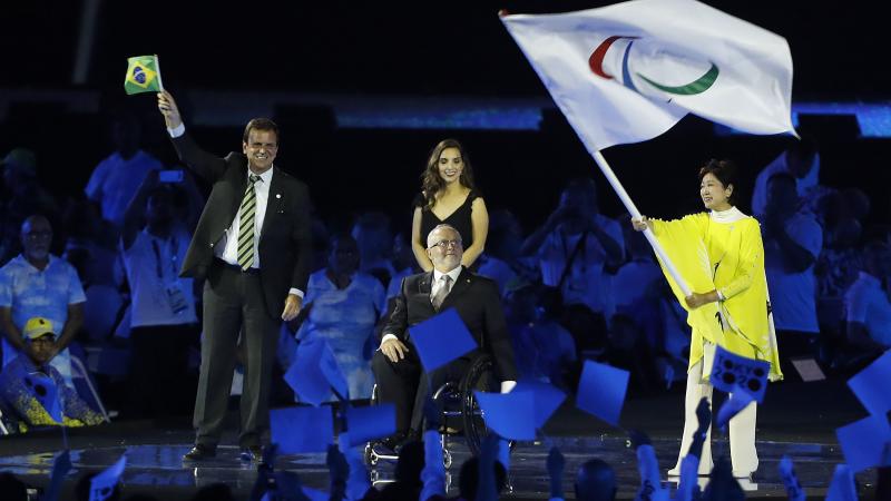 Three people on a stage, one woman waving a flag