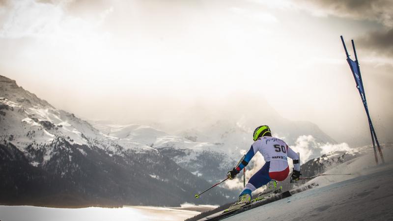 A standing alpine skiier goes down a slope with mountainous backdrop 