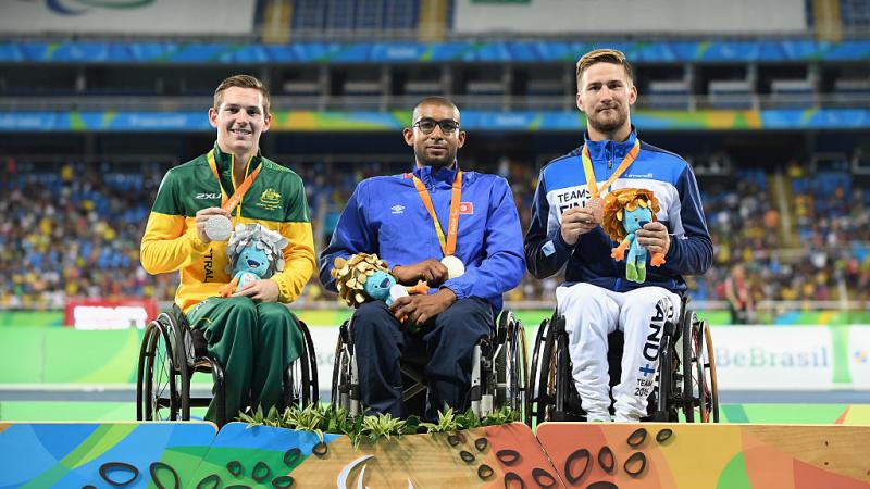 three men in wheelchairs smile holding their medals on the podium