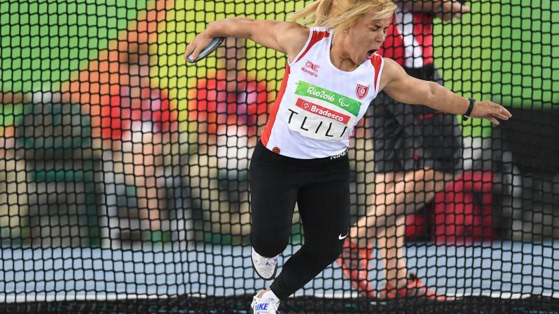 A woman in white vest and black bottoms pulls a face as she gets ready to throw a discus.