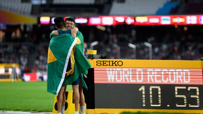 Two athletes hug on the track in front of a bulletin board showing 'WR'