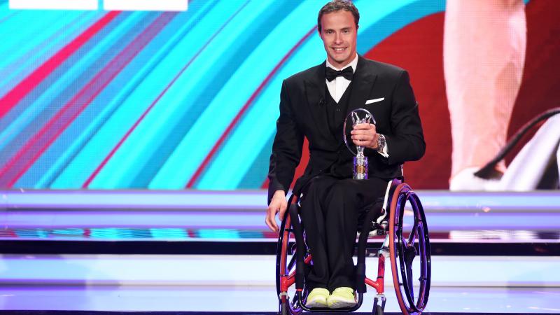 Man in wheelchair dressed in a suit holding a trophy