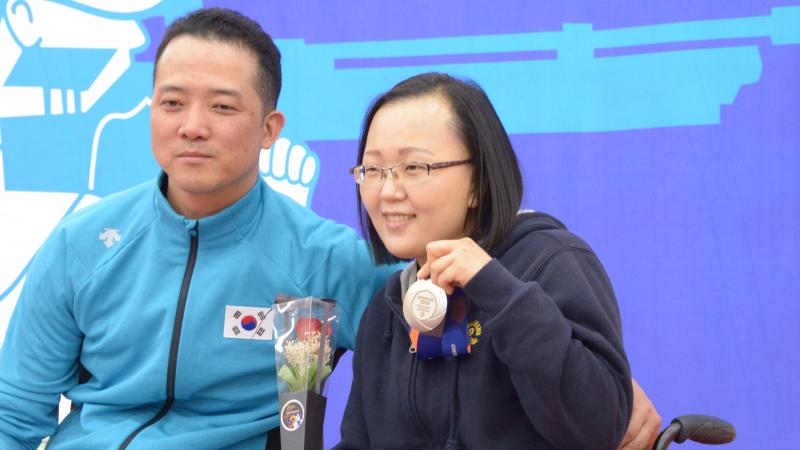 Man and woman in wheelchairs pose for photo with silver medal