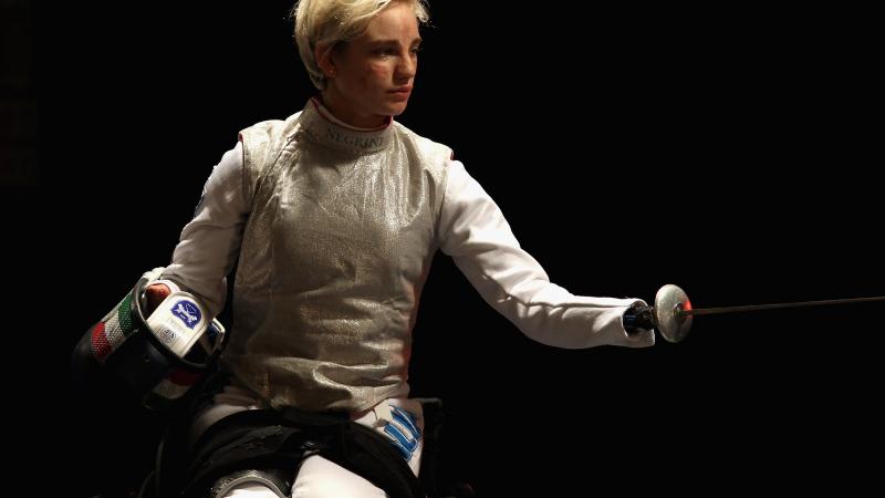 a female wheelchair fencer holds up her foil