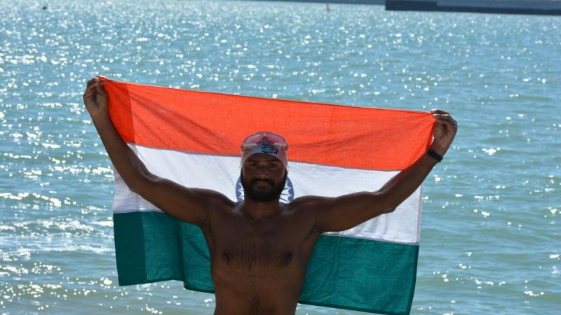 a male Para swimmer holds up in India flag beside the sea