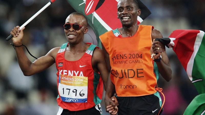 blind male runner Henry Wanyoike and his guide on a track carrying the Kenya flag