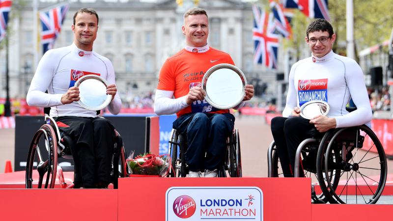 three male wheelchair racers including Marcel Hug and David Weir on a podium holding trophies