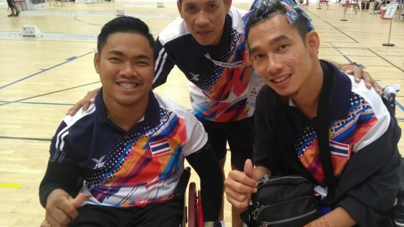two Thai boccia players and their coach standing behind them and smiling with his arms around their shoulders