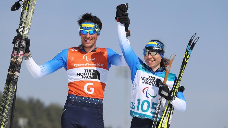 female Para Nordic skier Oksana Shyshkova holds hands with her guide as they raise their arms in victory