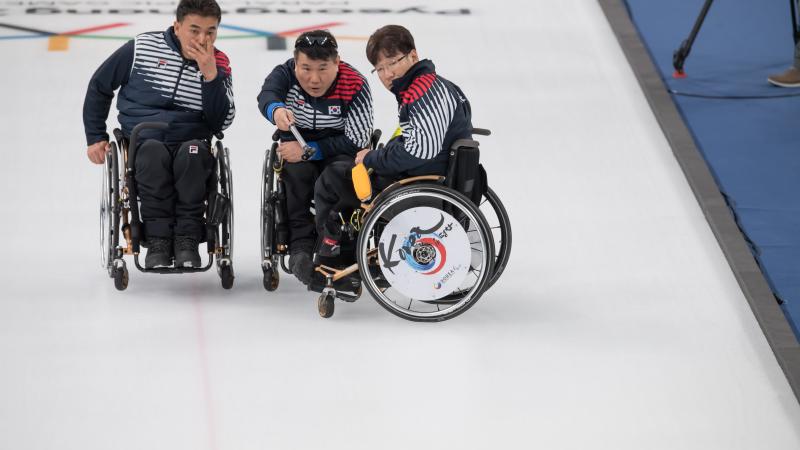 three male wheelchair curlers with Soon-Seok Seo on the right discussing how to play a stone