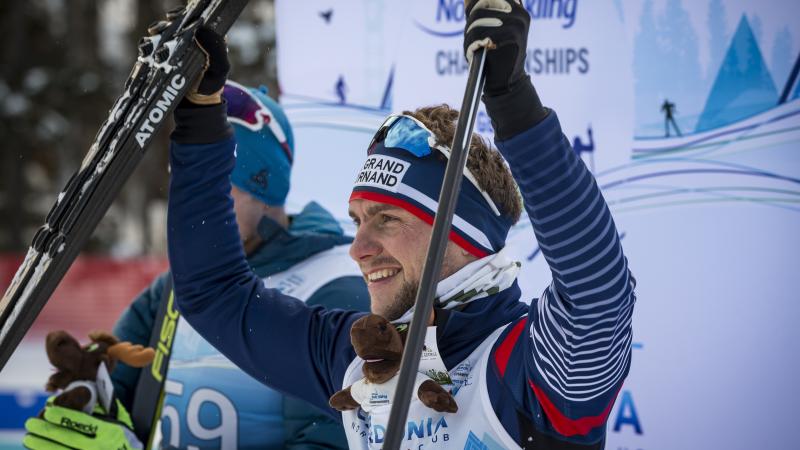 male Para Nordic skier Benjamin Daviet lifts up his skis in celebration on the podium