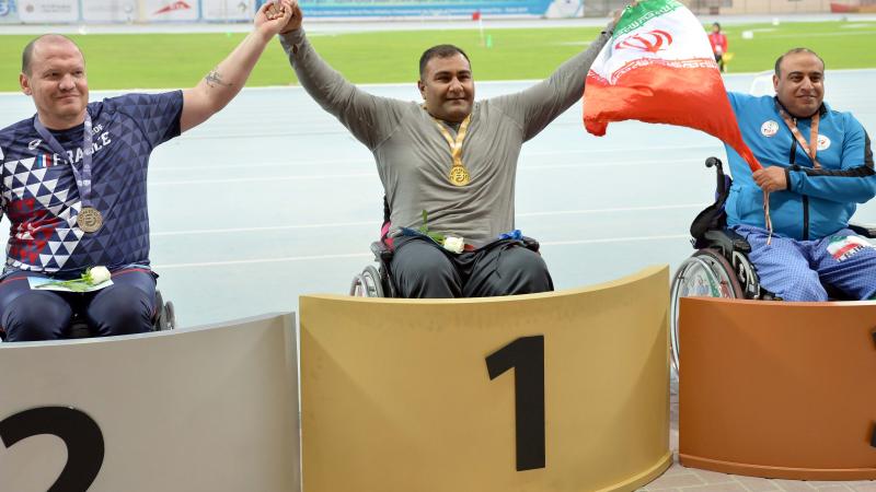 three male Para athletes in wheelchairs on the podium with Hamed Amiri in the centre holding an Iranian flag