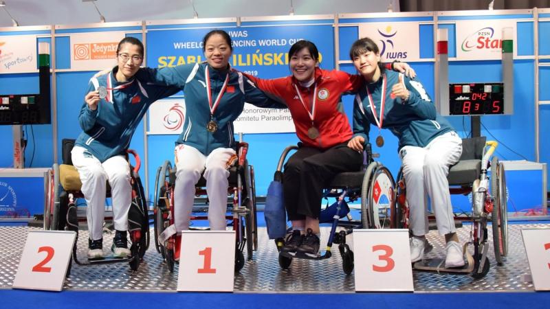 Four female wheelchair fencers put arms around each other and pose on a podium