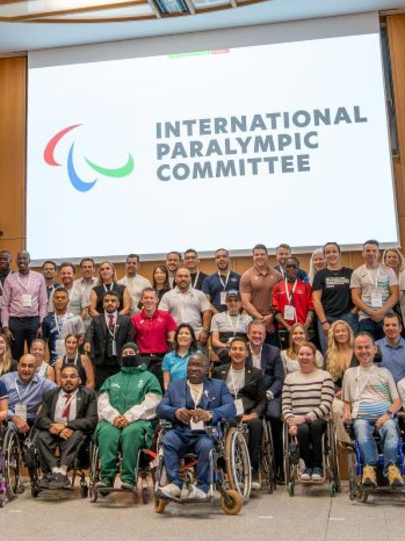 A group photo taken during the IPC Athletes' Forum - more than 70 athletes and officials are smiling in front of a screen with the IPC logo