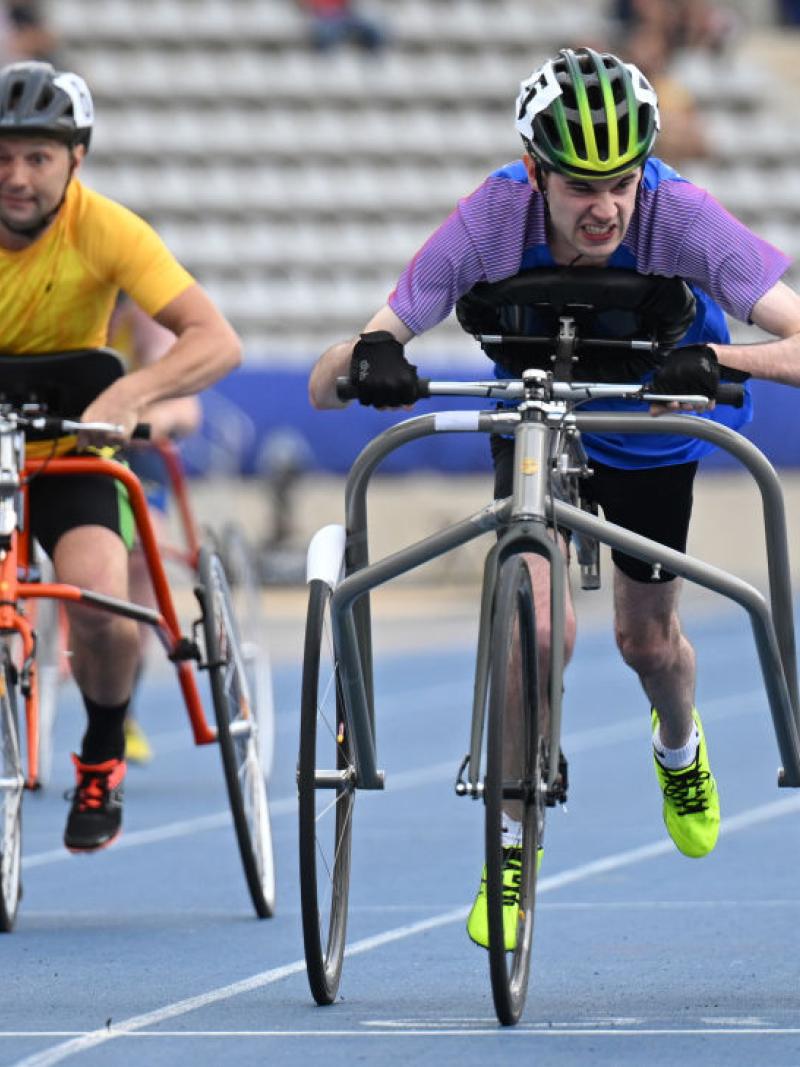 Two athletes are racing on a blue athletics track. They are running using a metal frame with three wheels