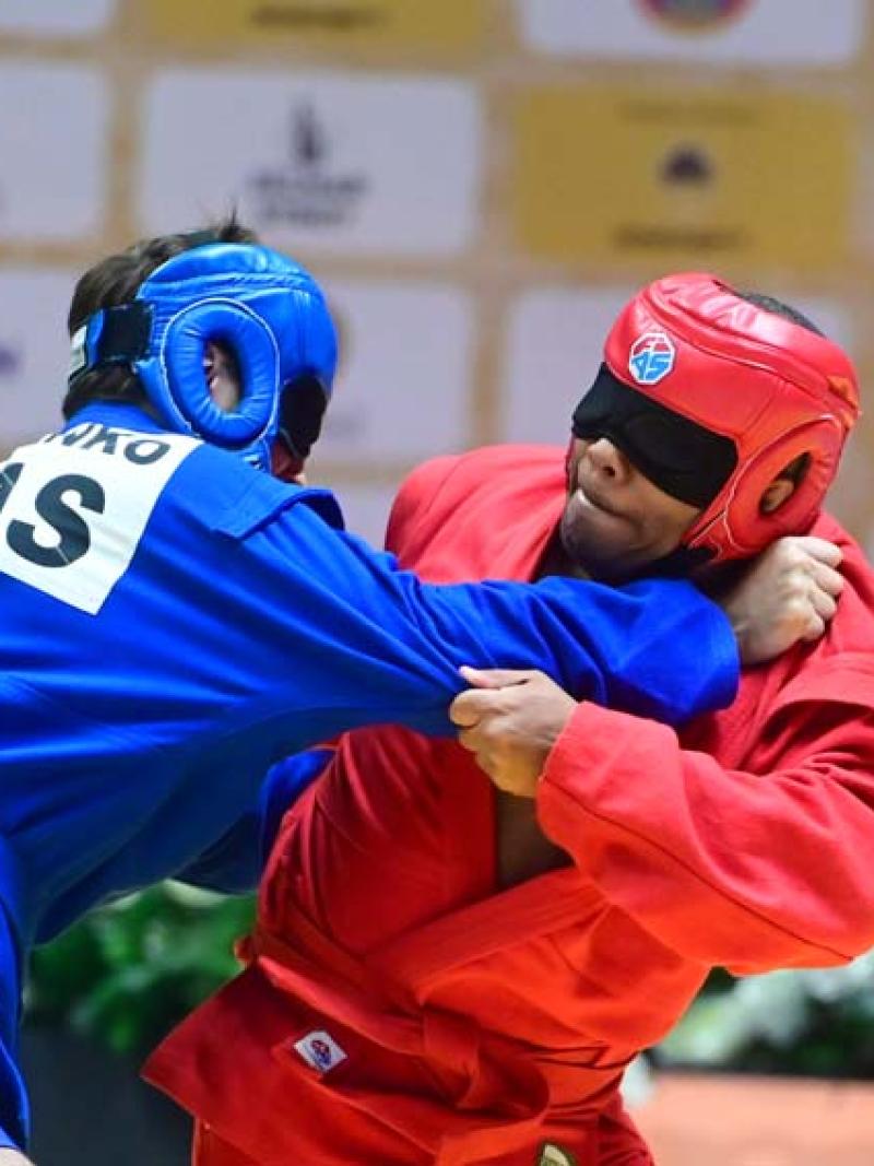 Two male athletes wearing blindfolds are wrestling - one athlete is wearing a red uniform and helmet while his opponent is wearing a blue helmet and gear