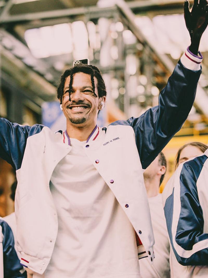 A male athlete wearing a white and blue hoodie is opening his arms wide to the crowd