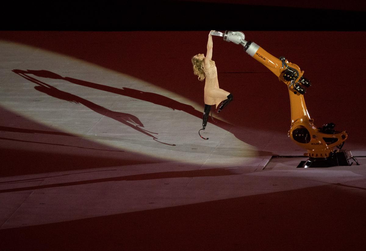 a female with prosthetic legs being raised into the air holding onto a robot arm