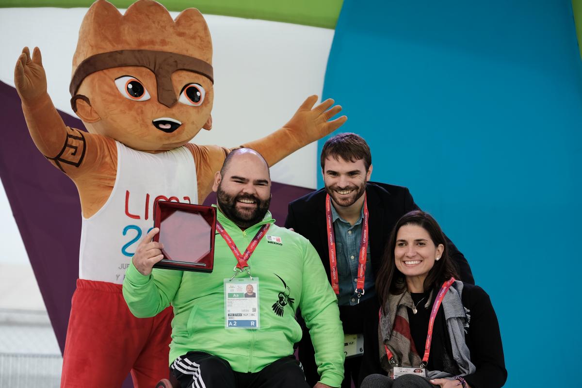 A male powerlifter holds up an award smiling with two other people and a furry mascot standing behind