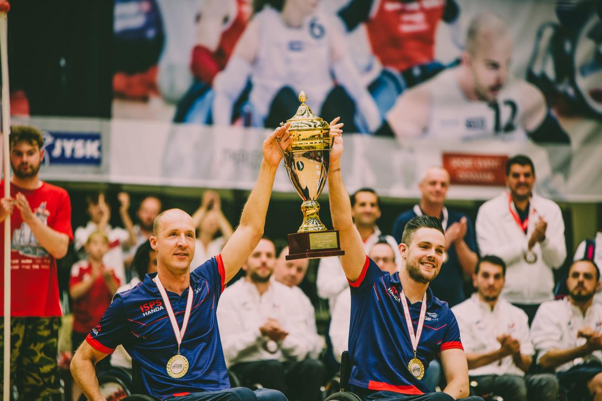 two male wheelchair rugby players with medals around their neck lift a gold cup into the air