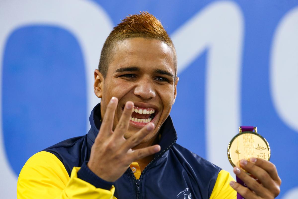 a male Para swimmer holds up four fingers and his gold medal