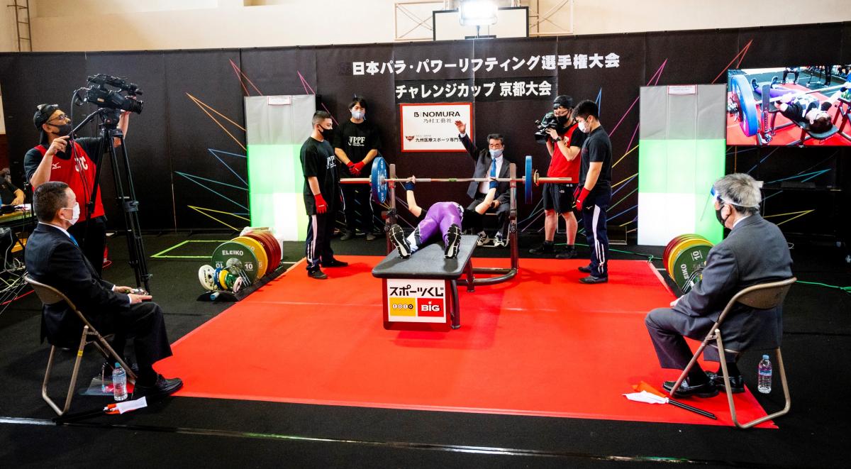 A person on a bench press during a Para powerlifting competition observed by three judges, three loaders and two cameramen