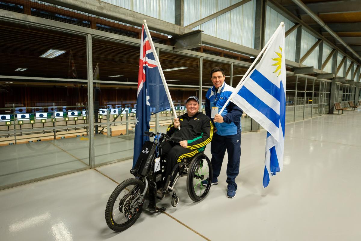 A man in a wheelchair holding the Australian flag posing next to a man standing and holding the flag of Uruguay