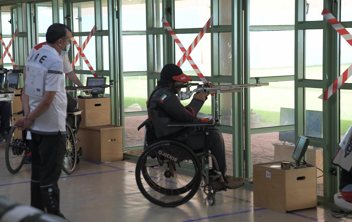 A man standing behind a man in a wheelchair shooting with a rifle in a shooting range