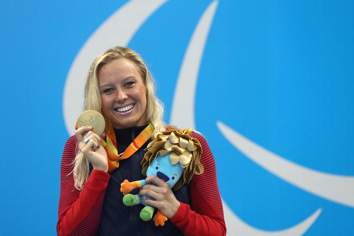 A woman posing for a photo with a smile on her face while holding a gold medal in her right hand and a mascot in her left hand