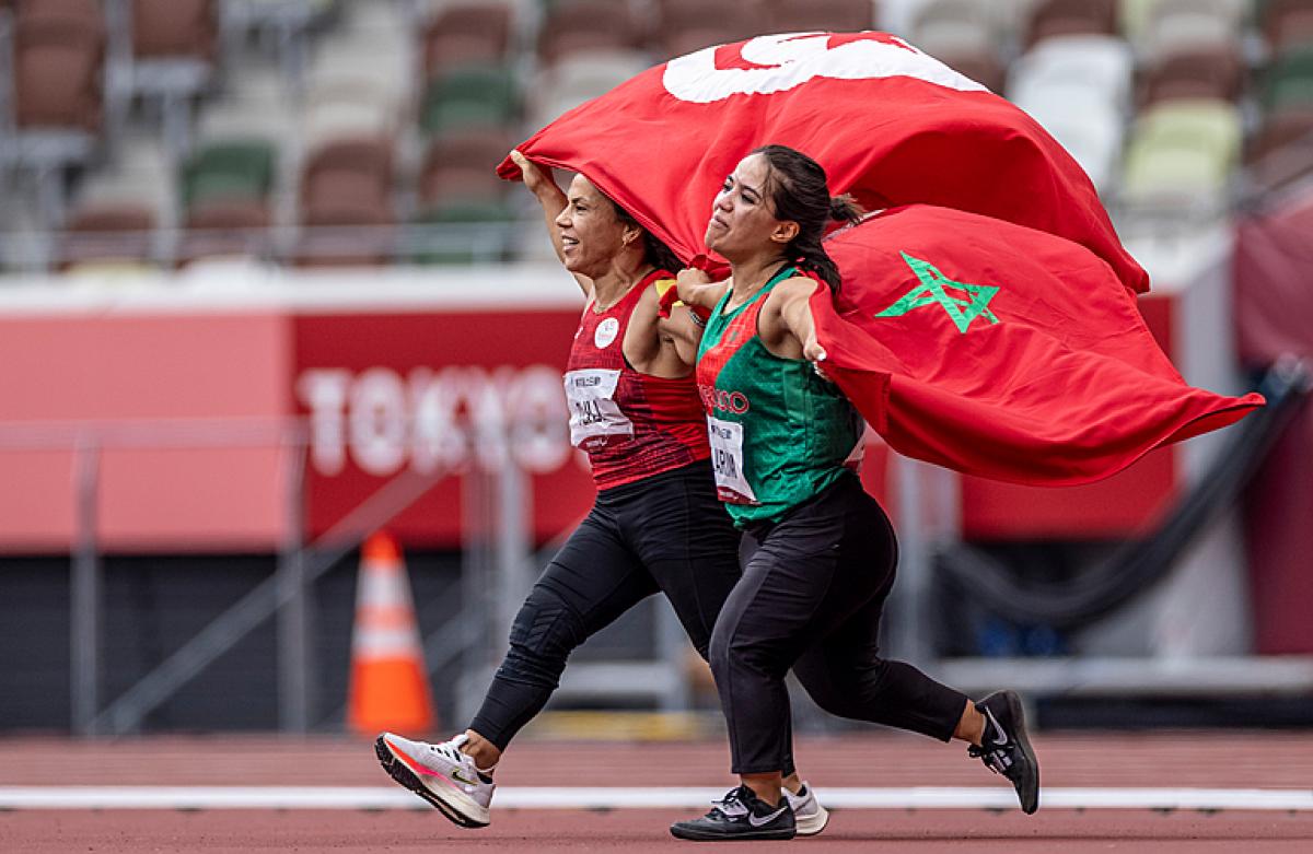 Two short stature women running on an athletics track holding the flags of Tunisia and Morocco