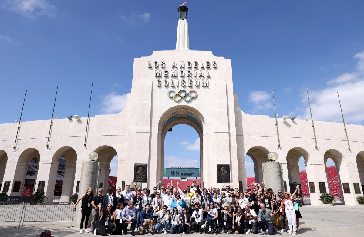 A group of people, many wearing LA28 shirts, pose for a photo in front of the Los Angeles Memorial Coliseum.