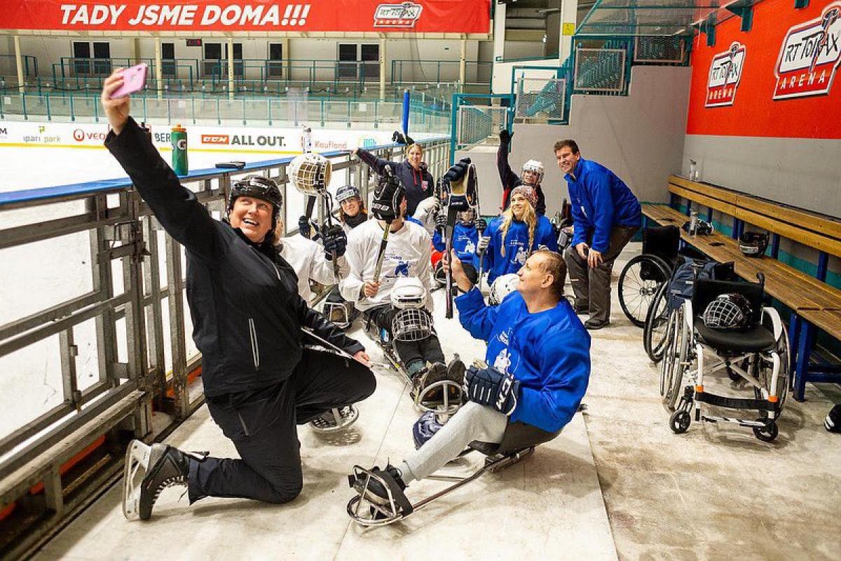 A group of 12 people posing for a selfie picture next to an ice hockey rink