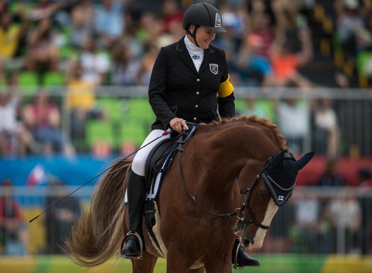 A female Para equestrian rider smiles as she rides a horse during competition.