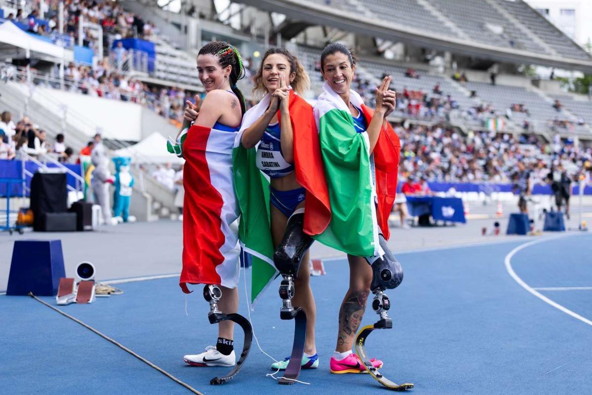 Three female athletes with prosthetic legs posing for a picture with the flag of Italy