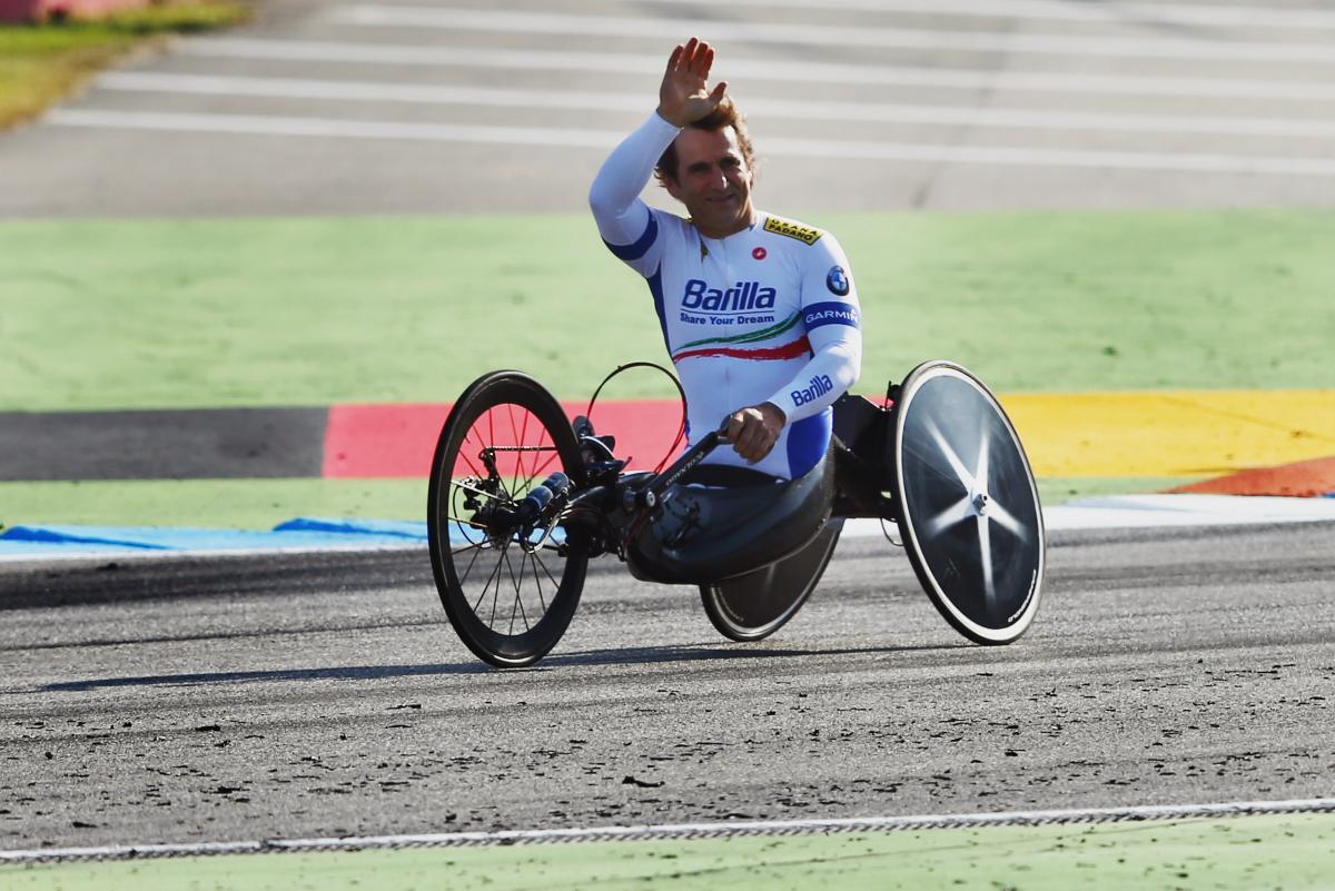 A man in a handcycle waves to spectators from a dirt track.