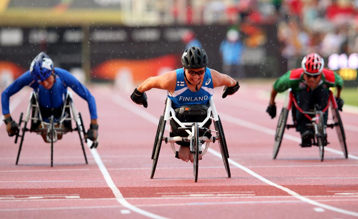 Three wheelchair racers approaching the finish line on the track of a stadium