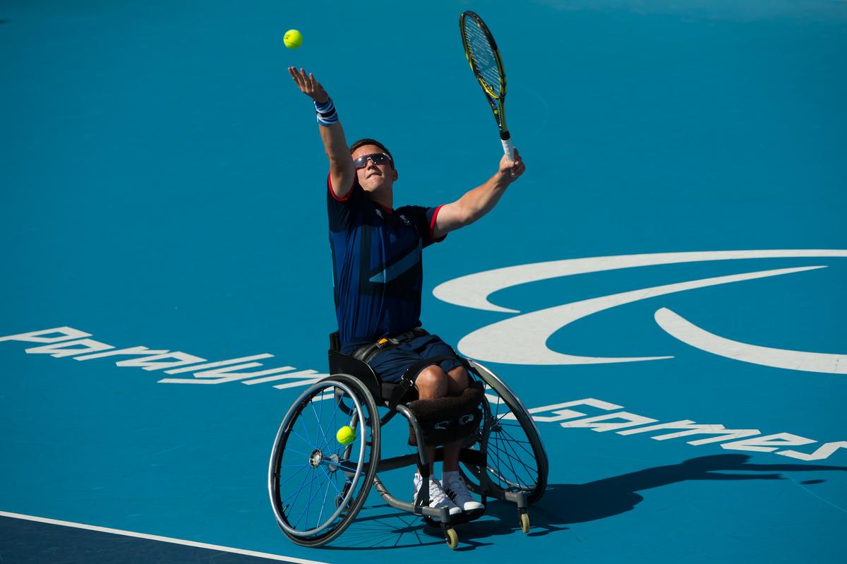 Man in wheelchair serves a ball on a tennis court.