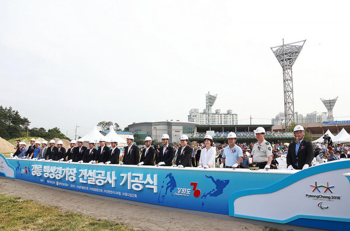 People in suits with construction helmets stand behind a banner in front of a construction area