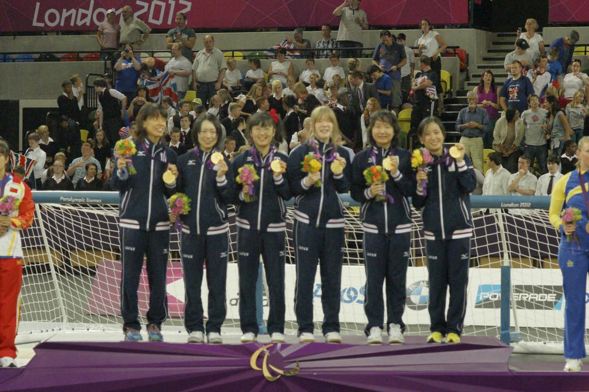 Six women in training suits on podium with medals and flower boquets.