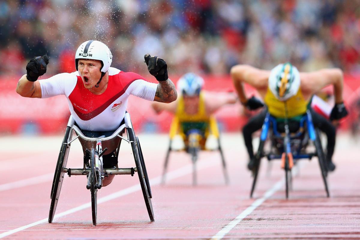 Man in racing wheelchair crosses finish line of a track throwing hands in the air, celebrating